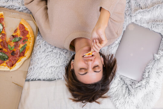 Woman east fast food from delivery on bed in bedroom at home at christmas new year time. Female alone enjoying fat food, pizza. Hungry for carbs.
 