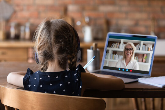 Rear View Close Up Little Girl Wearing Headphones Watching Webinar At Home, Using Laptop, Studying Online, Writing Notes, Student Listening To Older Woman Mentor Teacher, Homeschooling Concept