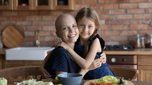 Head Shot Portrait Smiling Sick Mother Cancer Patient And Little Daughter Hugging, Standing In Kitchen, Cooking Salad Together, Unhealthy Hairless Mum And Adorable Child Girl Looking At Camera