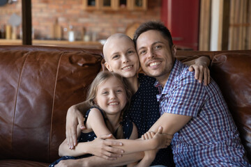 Fototapeta premium Portrait happy sick mother cancer patient, father and little daughter cuddling, looking at camera, smiling unhealthy woman and man with child posing for photo together, showing support and care