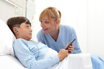 Happy child lying in bed in hospital room and smiling nurse using smartphone surfs the internet wearing earphones