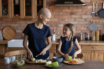 Happy sick hairless mother cancer patient and little daughter cooking together, chopping fresh...