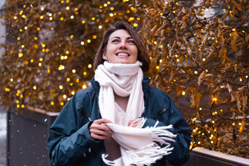 Caucasian happy smiling woman enjoying snow and winter, wearing warm scarf,  christmas lights on background, woman waiting for christmas miracle, street frame 2021    