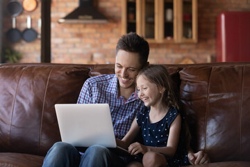 Laughing father and little daughter using laptop at home together, sitting on cozy couch in living room, happy dad and adorable child girl looking at screen, shopping, chatting online, watching video