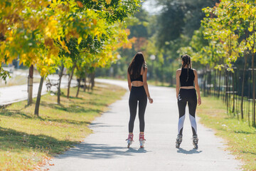 Rearview of two young fit women on roller skates riding outdoors on urban street in the park.