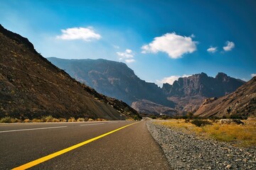 A landscape photo of the mountains, clouds, blue sky and large rocks taken from the middle of the road in Oman.