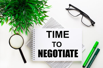 On a white background lies a notebook with the word TIME TO NEGOTIATE, glasses, a magnifying glass, green markers and a green plant