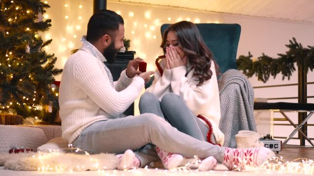 A marriage proposal on Christmas Eve. A young man gives a ring to his future bride while sitting on the floor near a tree. The young woman is surprised and very happy.