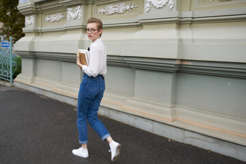 young woman in jeans and a shirt with a book in her hands near the building exterior education