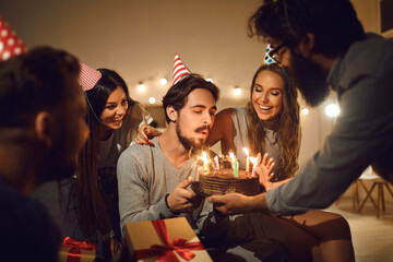 Young man in his 20s blowing candles on his birthday cake surrounded by happy friends