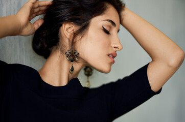 romantic brunette in an evening dress leaning against a gray wall indoors earrings makeup model