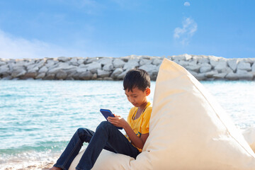 Asian boy sitting smile and playing mobile phone by the sea while traveling on the weekend.