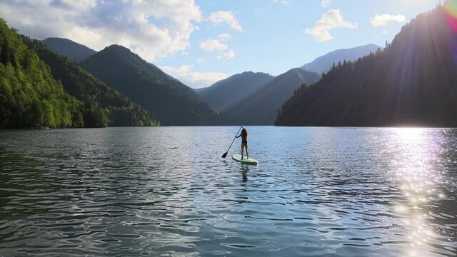 Concept of active tourism and travel. Aerial drone view of woman silhouette paddle on sup surfing board at mountain lake during sunset. Sporty girl doing water sport during summer holidays.