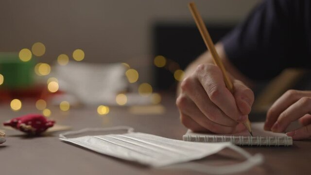 Slow motion orbit shot of closeup man writes Shopping list in notepad with a pencil under warm light in the evening with medicine mask near hand