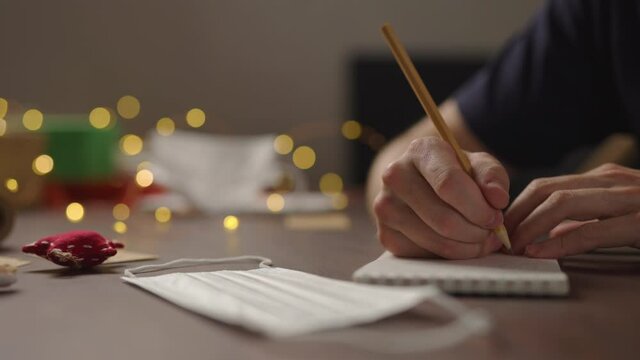 Slow motion orbit shot of closeup man writes Shopping list in notepad with a pencil under warm light in the evening with medicine mask near hand