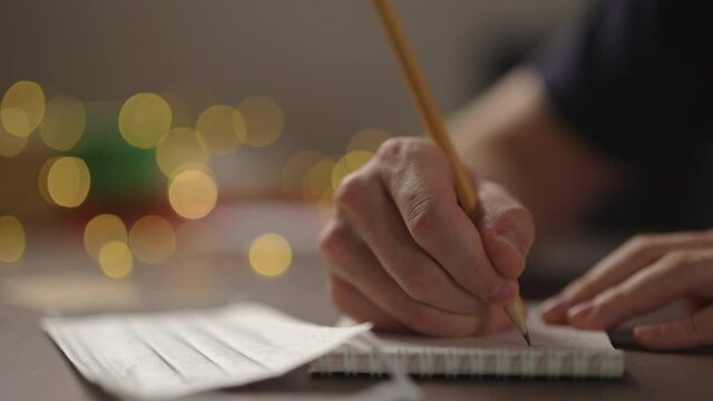 Slow motion closeup man writes Shopping list in notepad with a pencil under warm light in the evening with medicine mask near hand