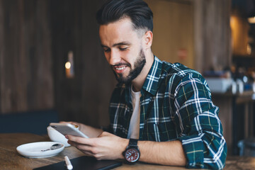 Happy Caucaisan male blogger reading funny text publication and smiling during coffee time in cafeteria, young hipster guy connecting to public wifi internet on modern smartphone technology