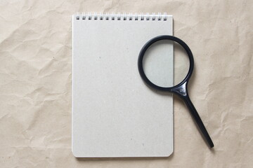 Fototapeta premium Gray notepad with white coiled spring and magnifieron a background of beige crumpled craft paper