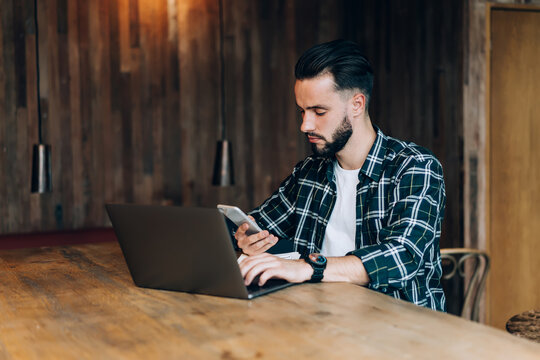 Millennial blogger in shirt using bluetooth connection on modern smartphone and laptop technologies for sharing media files during freelance remote working, man checking received cellular notification