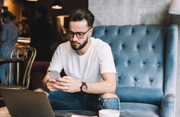 Millennial blogger in spectacles using bluetooth connection on smartphone and laptop technologies...