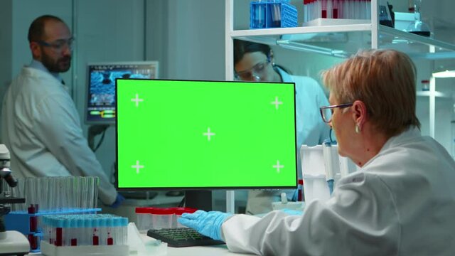 Senior doctor working at computer with green screen in modern equipped lab late at night. Team of microbiologists doing vaccine research writing on device with chroma key, isolated, mockup display.