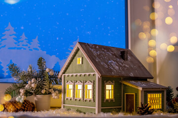 On windowsill, among Christmas tree decorations, night light in form of village house glows, against background of night snow window and paper decorations.