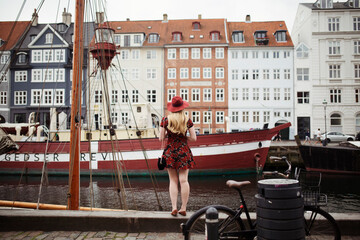woman with long blonde hair and sun hat in copenhagen in front of colorful buildings traveling on...