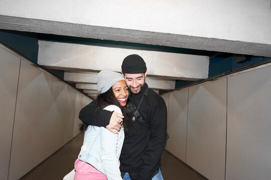 Life flash style photo-Young interracial couple of lovers wearing face masks in an underground subway corridor in each other's arms down an old public transportation corridor enjoying their love.