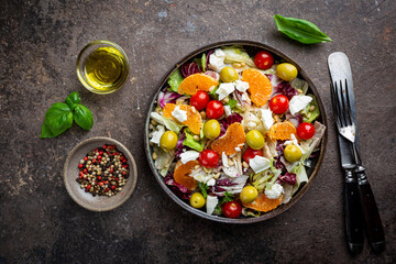 Fresh green mixed salad leaves frisee, radicchio and lamb's lettuce with tomato, olives and tangerine in a plate, top view.
