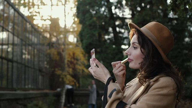 Beautiful woman in a hat and coat standing in an autumn park and applying lipstick, looking in a pocket mirror in a nature garden, slow motion