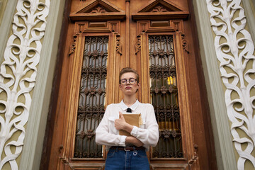 female student with glasses with a book in hands near the high door of the institute of education building