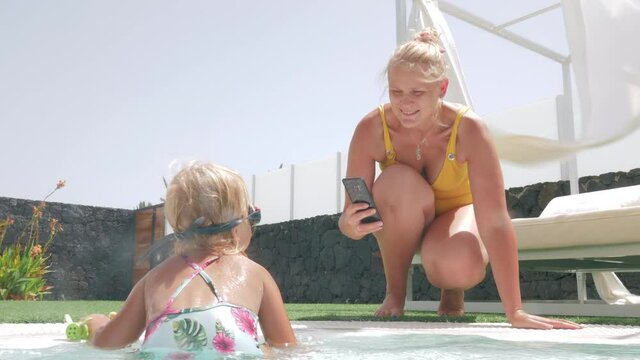 Mother taking summer pictures of little daughter swimming in the pool. Capturing happy childhood