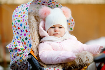 Sad crying hungry baby girl sitting in the pram or stroller on cold autumn, winter or spring day. Weeping child in warm clothes, fashion stylish baby coat and hat. Snow falling down