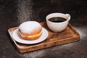 Hot coffee in white cup and fresh powdered sugar donut on a wooden tray. Low key. Black background. Breakfast concept
