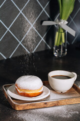 Hot coffee in white cup and fresh powdered sugar donut on a wooden tray. Low key. Black background. Breakfast concept