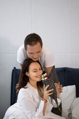 Beautiful couple in love - a man and a woman of about 40, dressed in white in bed with white sheets. girl holds a bouquet of roses, smiles and glows with happiness. Family morning tender moment