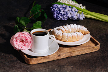 Hot coffee in white cup, powdered croissant and flowers blue hyacinth, pink rose on wooden tray. Coffee, croissant and flowers in low key on the dark background. 8 March concept