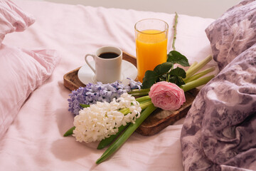 Breakfast in bed with hot coffee, orange juice and fresh flowers rose and hyacinth on a wooden tray in pink-purple bedroom. Breakfast concept