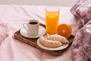 Breakfast in bed with hot coffee, powdered croissant, orange juice on a wooden tray in pink-purple bedroom. Breakfast concept