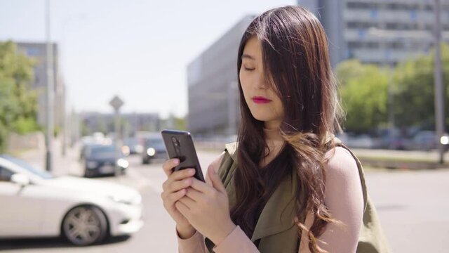 A young Asian woman works on a smartphone in a street in an urban area - cars drive on the busy road in the blurry background