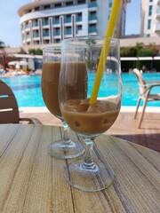Glass transparent glasses with cold coffee and a straw on the table by the pool