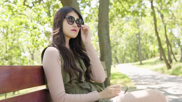 A young Asian woman puts on sunglasses and relaxes as she sits on a bench in a park on a sunny day