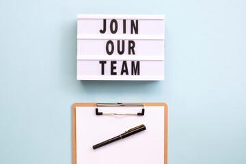 'Join our team' words on a lightbox, clipboard with blank sheet of paper on blue background. Overhead, from above, flat lay. Copy space.