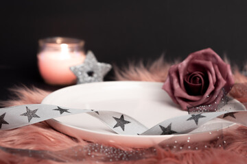 Empty plate, ribbon with stars, glitter star, burning candle, soft focus background, pink rose, black background, fluffy pink table cloth