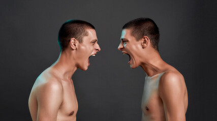 Fototapeta na wymiar Two young half naked angry caucasian twin brothers arguing, shouting while standing face to face isolated over dark grey background