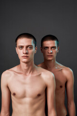 Vertical shot of two young caucasian half naked twin brothers looking at camera while posing in studio, standing isolated over grey background
