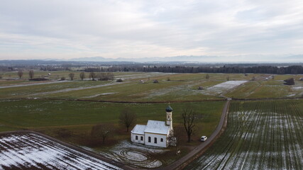 Raisting, Germany Bavaria - December 12, 2020: St. Johannes d. Täufe small white snowy roof wedding Chapel or Church sits in the country farm fields with winter trees. Near Munich and the Alps.
