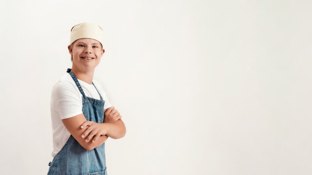 Disabled boy with Down syndrome dressed as a cook in apron and hat smiling at camera while standing with arms crossed isolated over white background