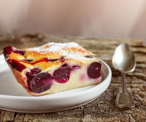 A slice of clafoutis cherry pie in a gray plate and a spoon on the old wooden table close-up