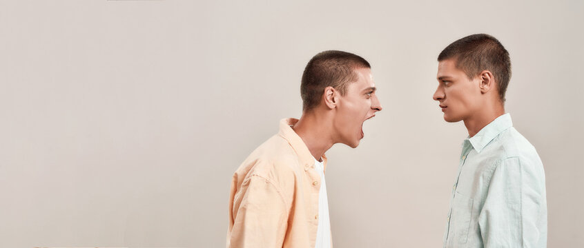 Portrait of two young caucasian men, twin brothers arguing, shouting while standing face to face isolated over beige background, panoramic banner